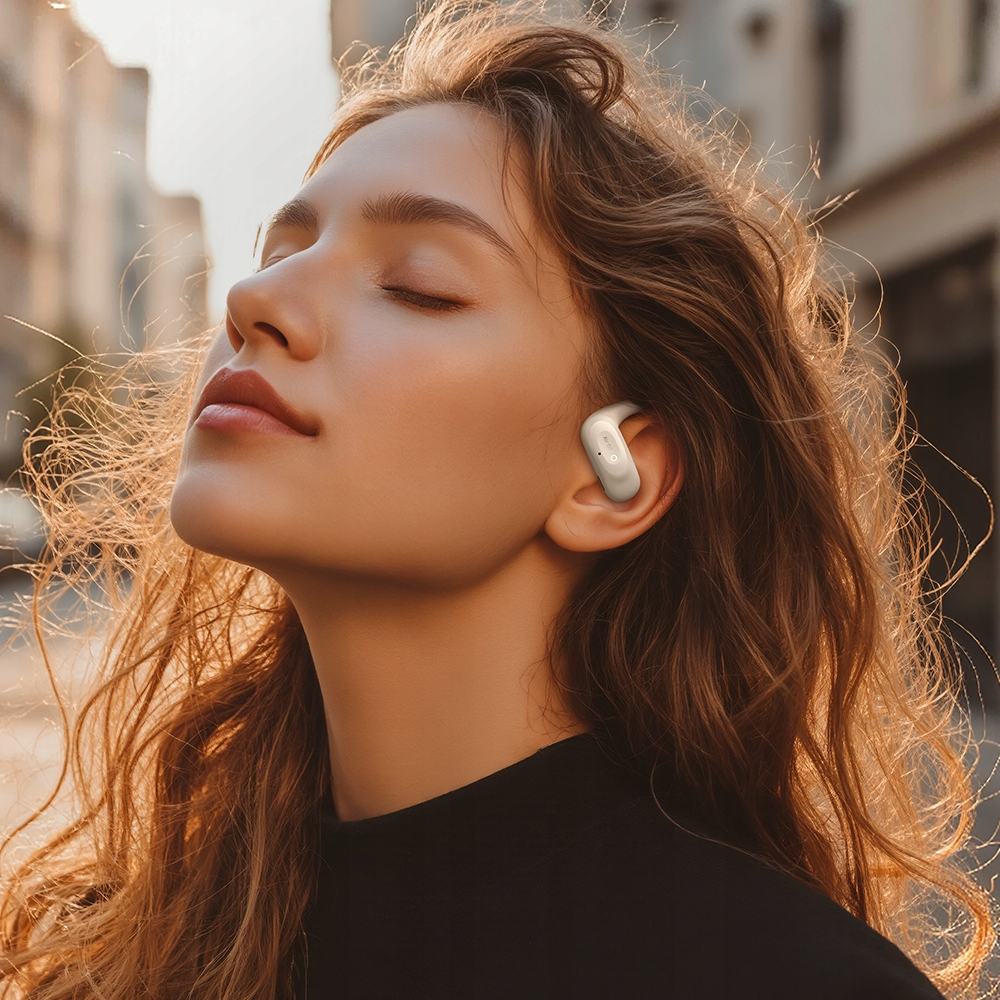 Una donna con gli occhi chiusi e i capelli castani al vento indossa un auricolare bianco all'aperto.