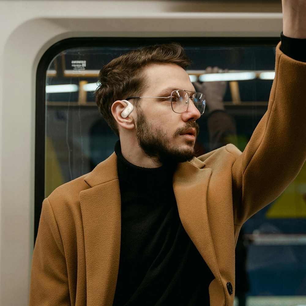 Un uomo con gli occhiali e la barba indossa un cappotto marrone e abiti neri in metropolitana.