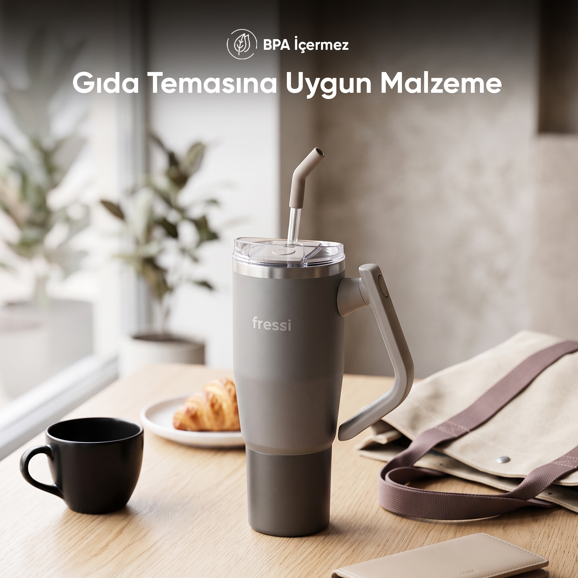 Ahşap masada pipetli gri Fressi bardak, yanında fincan, kruvasan ve çanta.