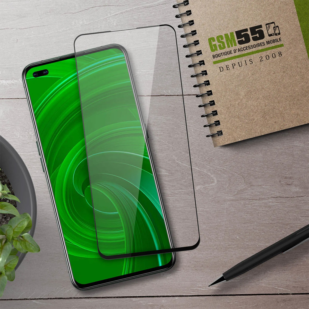 Smartphone con display verde, pellicola protettiva in vetro, taccuino e penna su tavolo in legno.