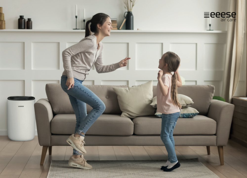 Mutter und Tochter tanzen fröhlich im Wohnzimmer vor einem Sofa, links steht ein weißer Luftentfeuchter.