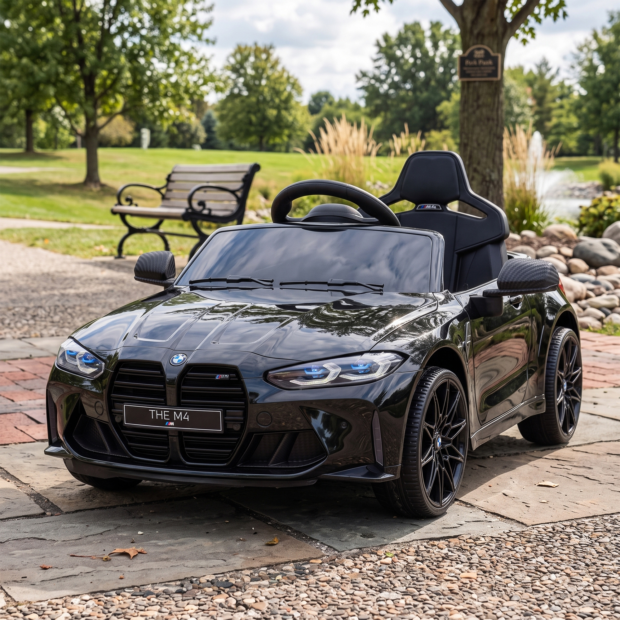 Un coche de juguete BMW M4 negro aparcado en un sendero de piedra en un parque, con un banco y árboles al fondo.