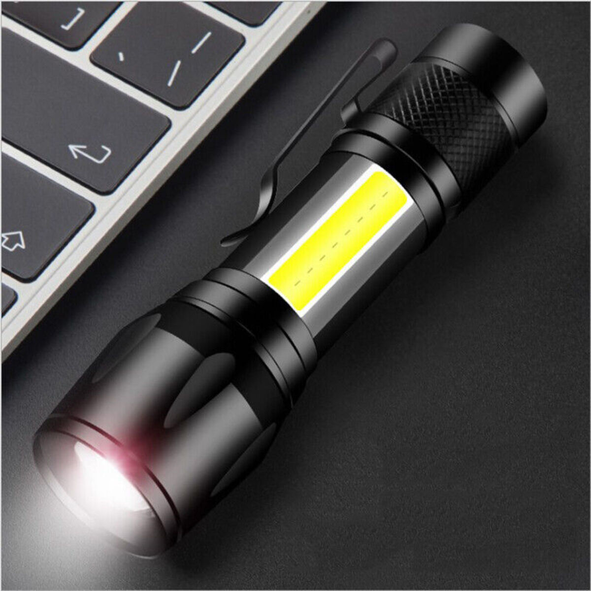 Eine kleine schwarze LED-Taschenlampe mit Clip liegt auf einer dunklen Oberfläche neben einer Tastatur.