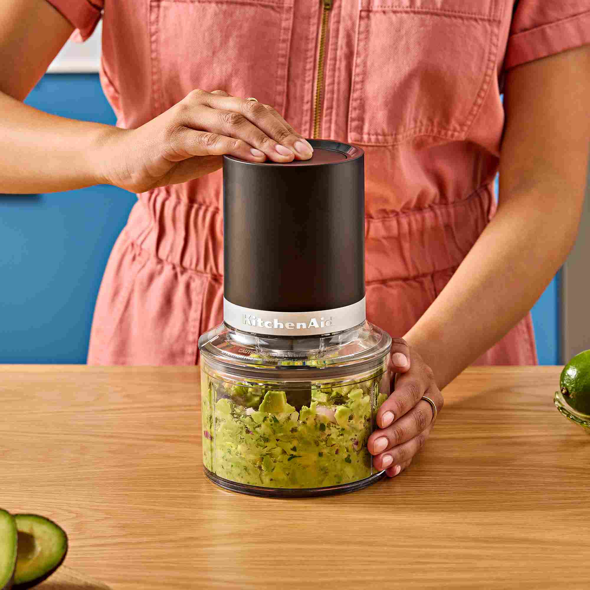 Eine Person nutzt einen schwarzen KitchenAid-Zerkleinerer auf einem Holztisch, um Guacamole zuzubereiten.