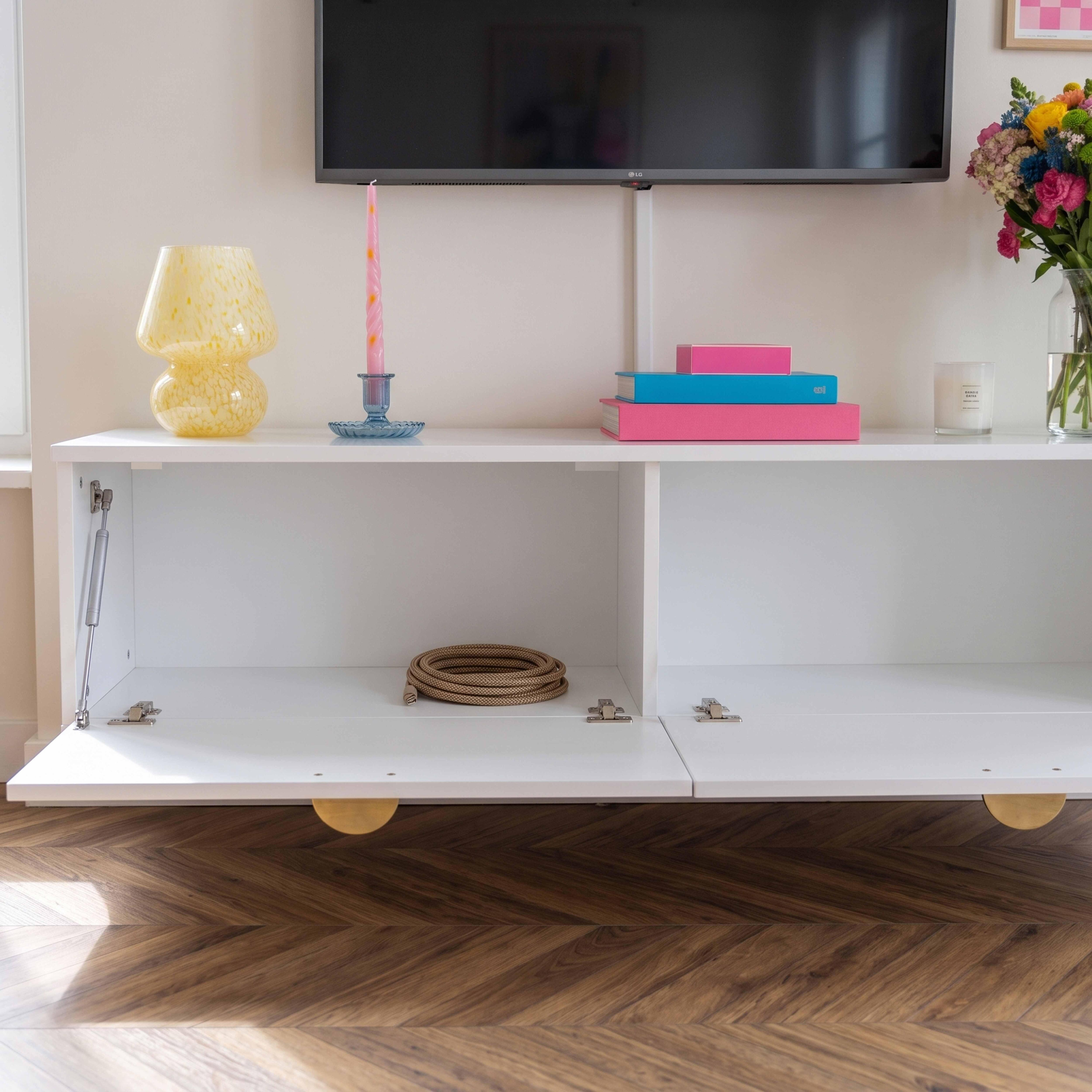 Ein weißes Sideboard unter einem Fernseher mit bunten Dekorationen oben drauf und einer aufgerollten Schnur drinnen.