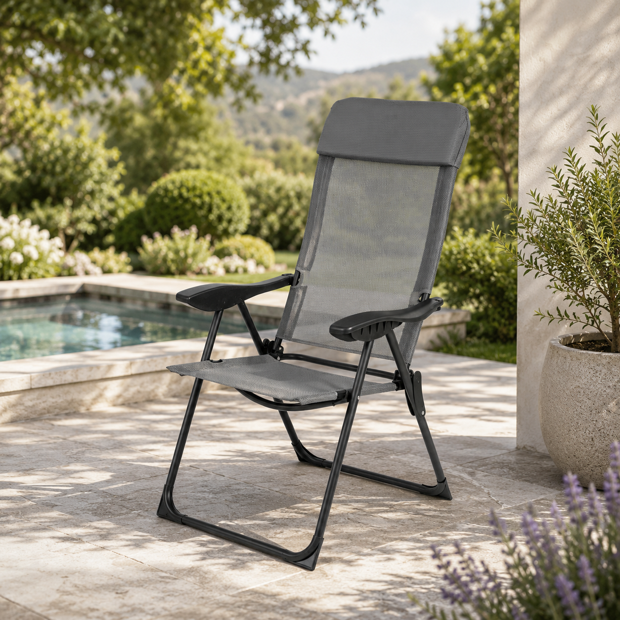 Una silla plegable gris en un patio de piedra junto a la piscina, rodeada de jardín.