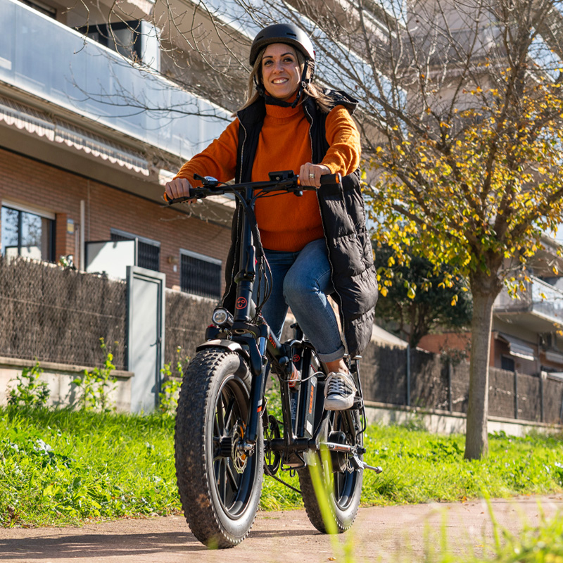 Una mujer con casco y jersey naranja monta en bicicleta eléctrica en un camino.