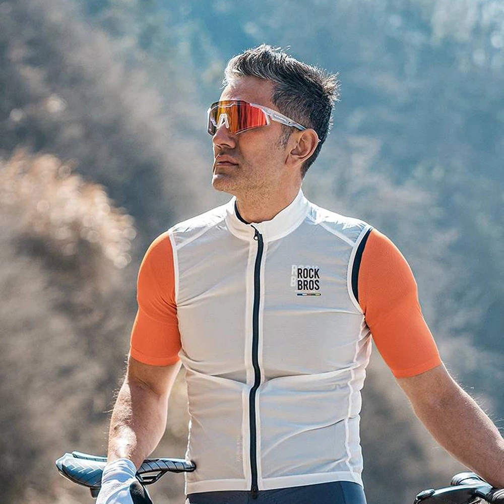 Un ciclista con gilet bianco, maniche arancioni e occhiali rossi, posa all'aperto con una bicicletta.