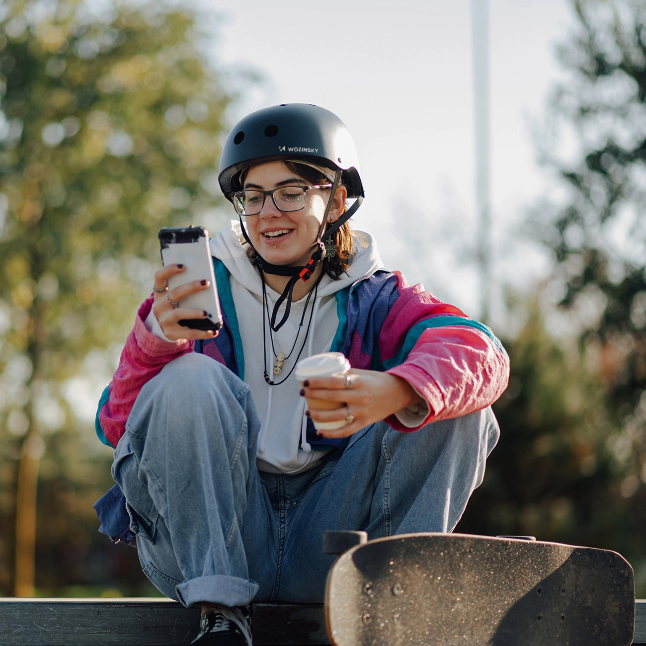 Una skater con casco e occhiali siede, guarda lo smartphone e tiene una tazza di caffè.