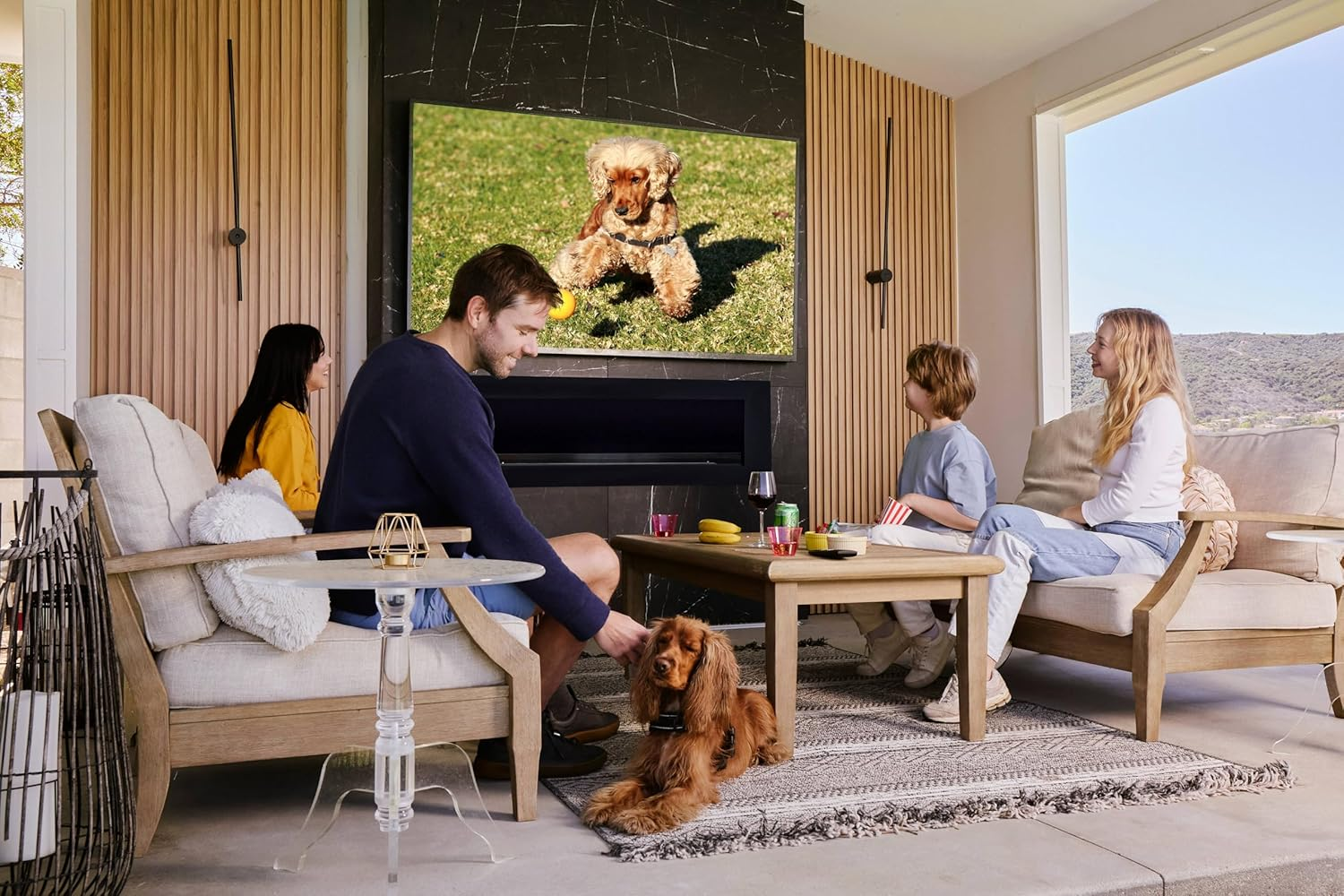 Eine Familie sitzt im Wohnzimmer vor einem Fernseher mit einem Hundebild, während ein echter Hund am Boden liegt.