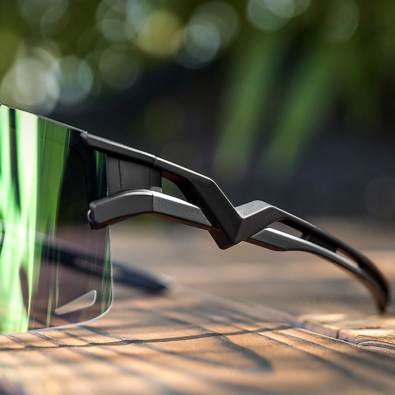 Primo piano di moderni occhiali da sole sportivi neri con lente verde riflettente su legno.