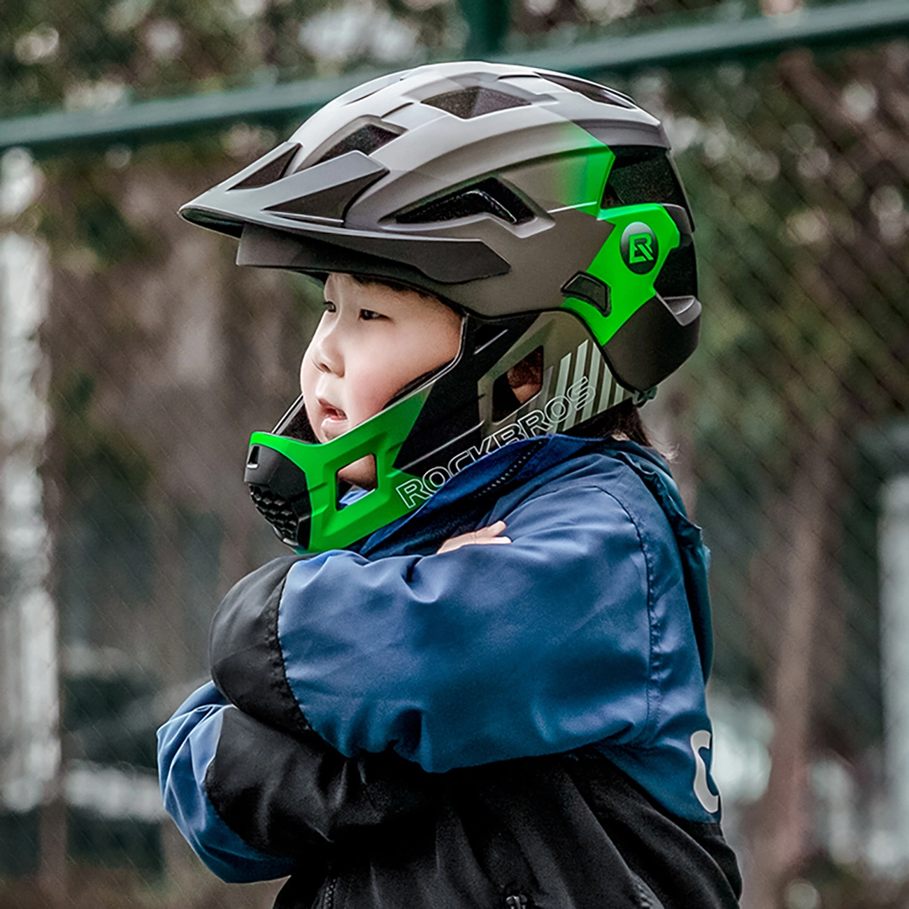 Un bambino con un casco da bici nero e verde e una giacca blu e nera si trova davanti a una recinzione.