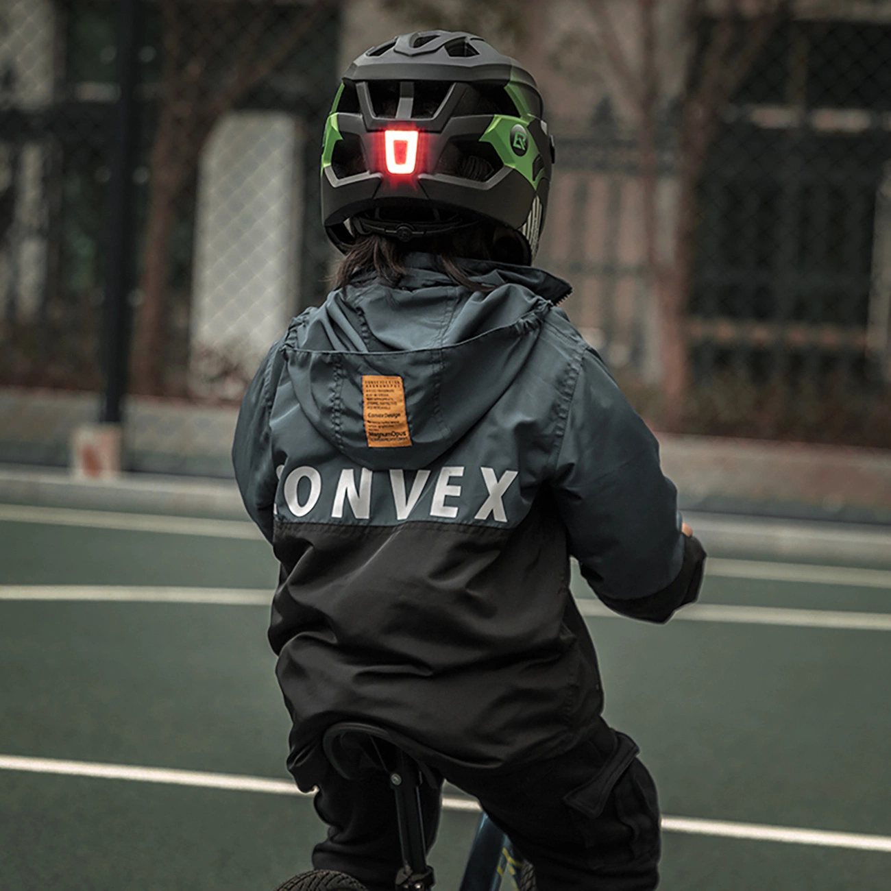 Un bambino con casco nero e verde con luce rossa e giacca scura con CONVEX sul retro, in bicicletta.