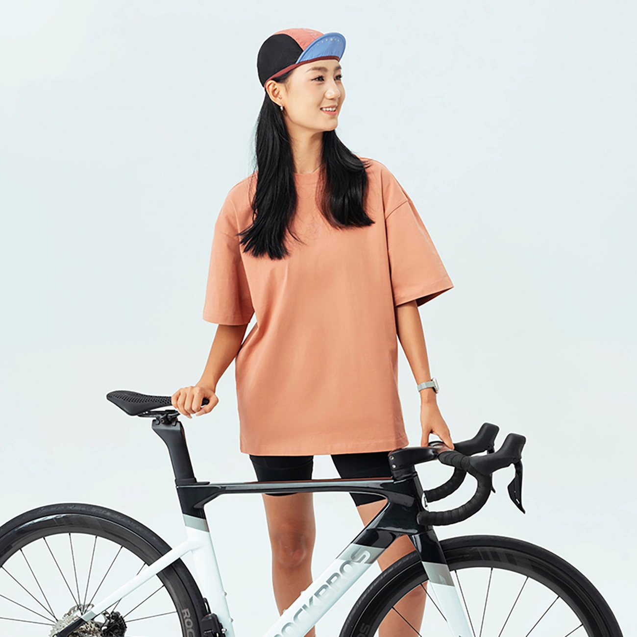 Una giovane donna con capelli neri e berretto colorato sorride accanto a una bici da corsa su sfondo bianco.