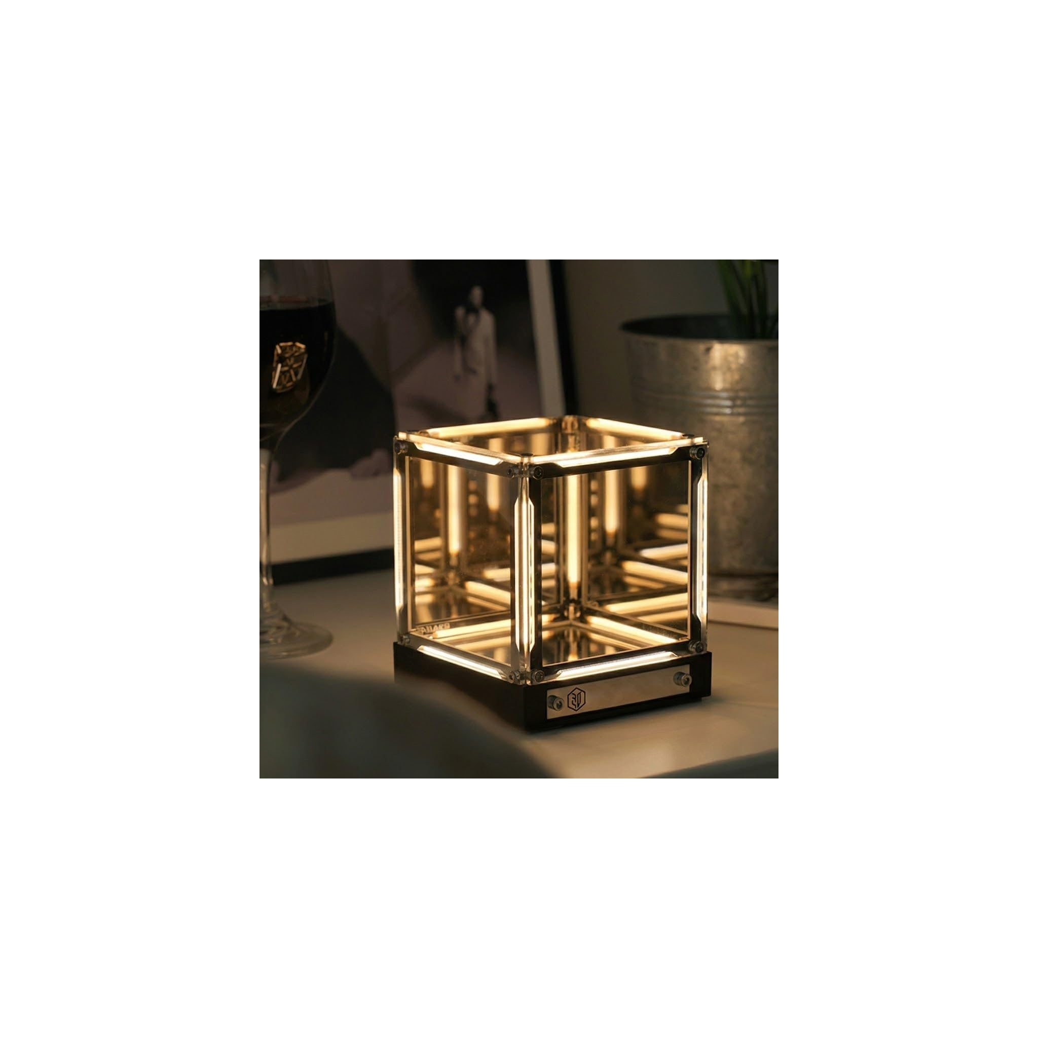 Eine Leuchtende Würfellampe auf einem Tisch mit Weinglas und Metalltopf im Hintergrund.
