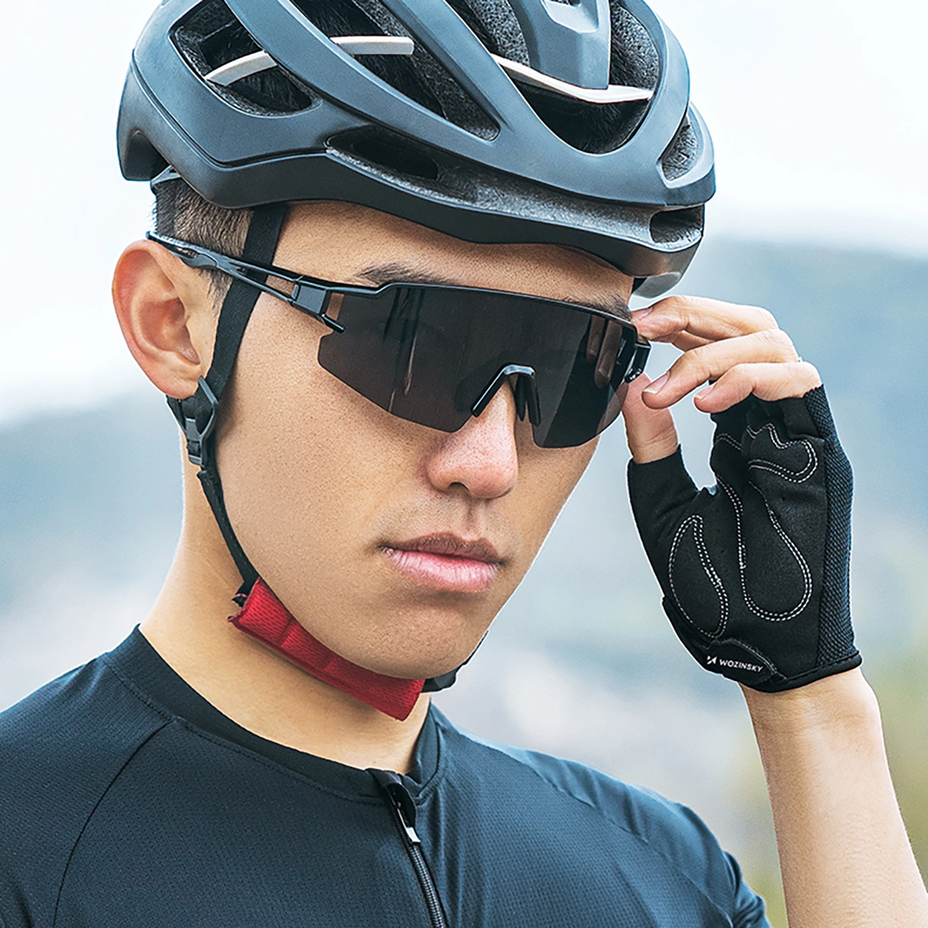 Un ciclista con casco nero, occhiali da sole sportivi scuri e guanti da ciclismo che si sistema gli occhiali.