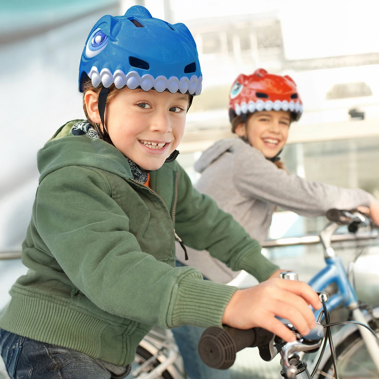Due bambini sorridenti vanno in bicicletta indossando caschi a forma di dinosauro, uno blu, l'altro rosso.