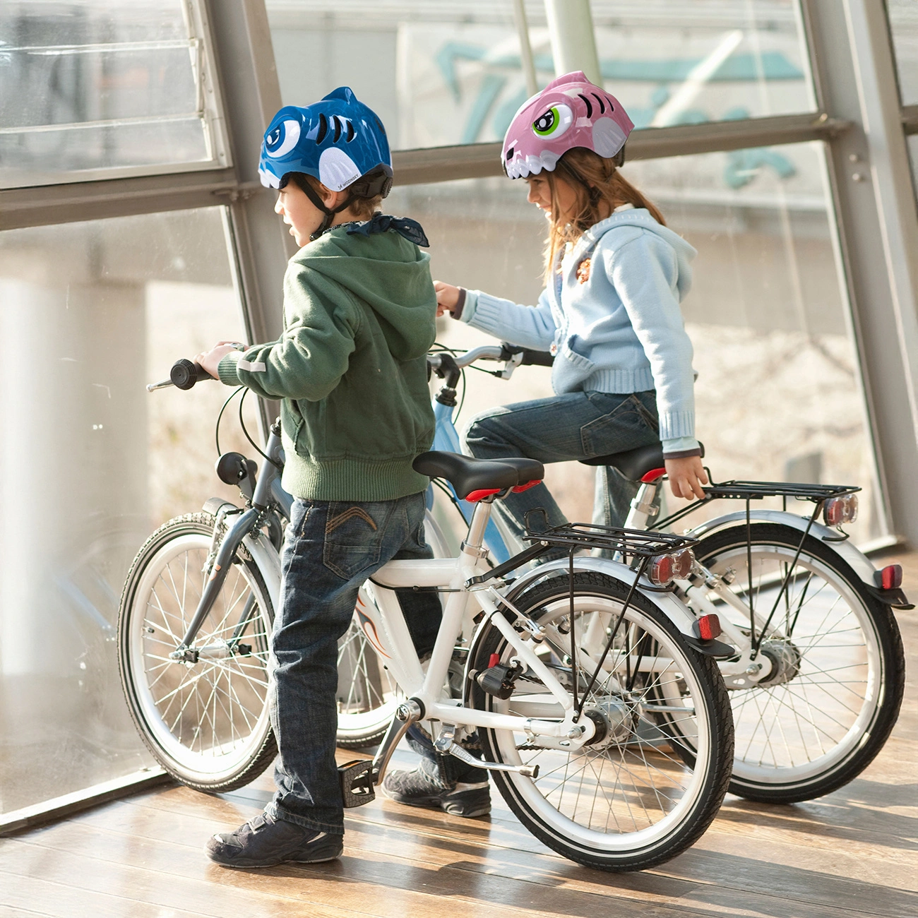 Due bambini con caschi colorati sono seduti sulle biciclette davanti a una grande finestra.