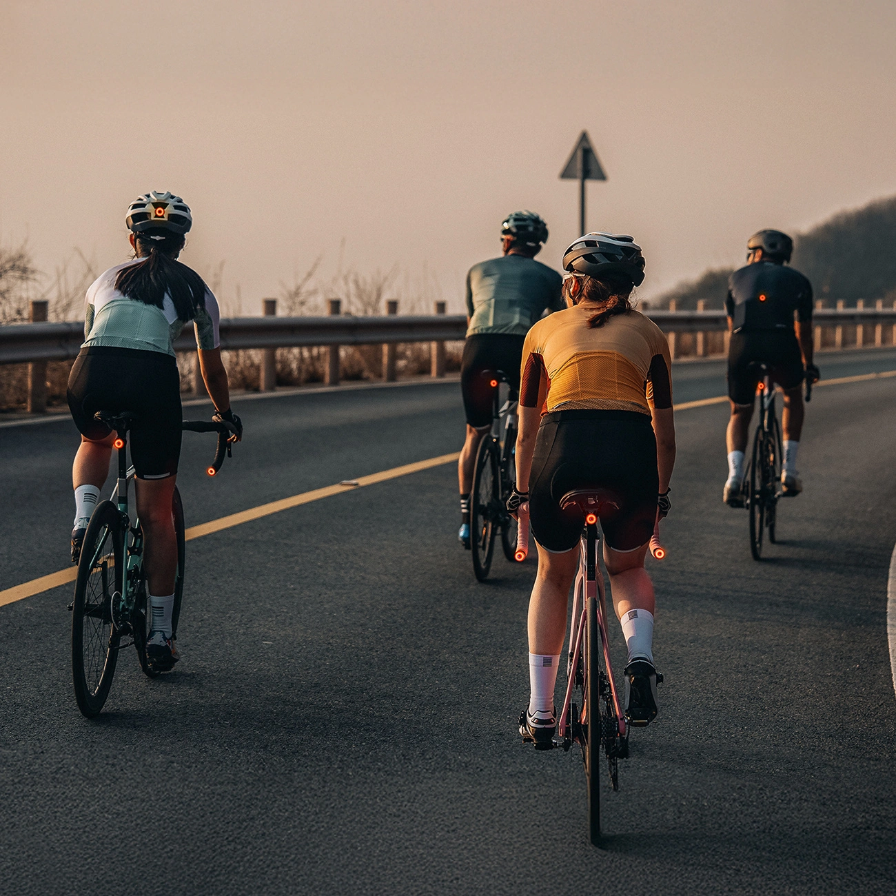 Quattro ciclisti in abbigliamento sportivo e caschi pedalano su una strada asfaltata verso l'orizzonte.