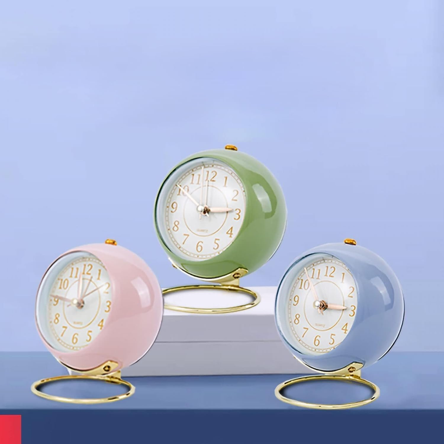 Drei runde Wecker in Rosa, Grün und Blau mit goldenen Ständern stehen vor einem hellblauen Hintergrund.