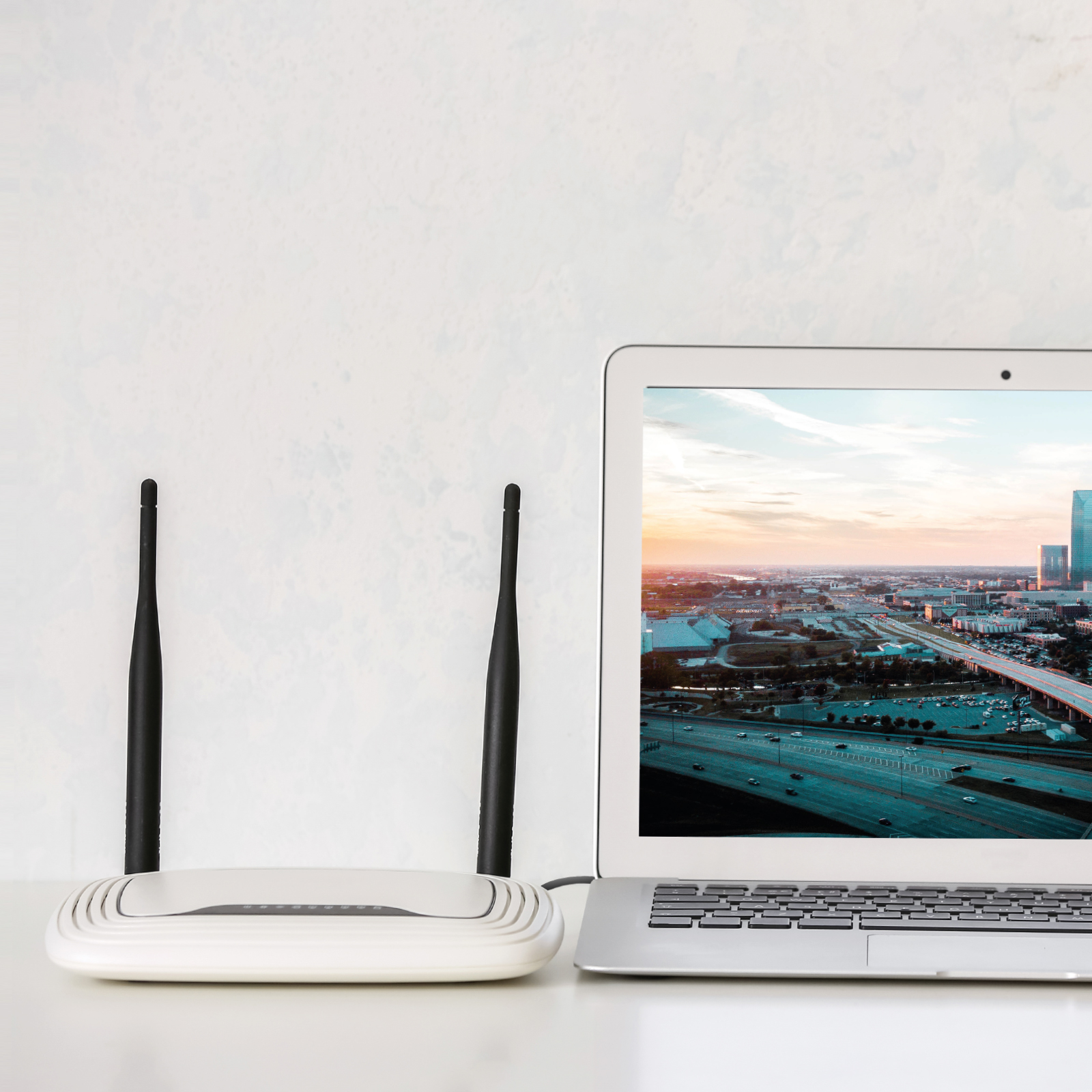 Ein weißer WLAN-Router mit zwei Antennen steht auf einem Tisch neben einem Laptop mit einer Stadtansicht auf dem Bildschirm.