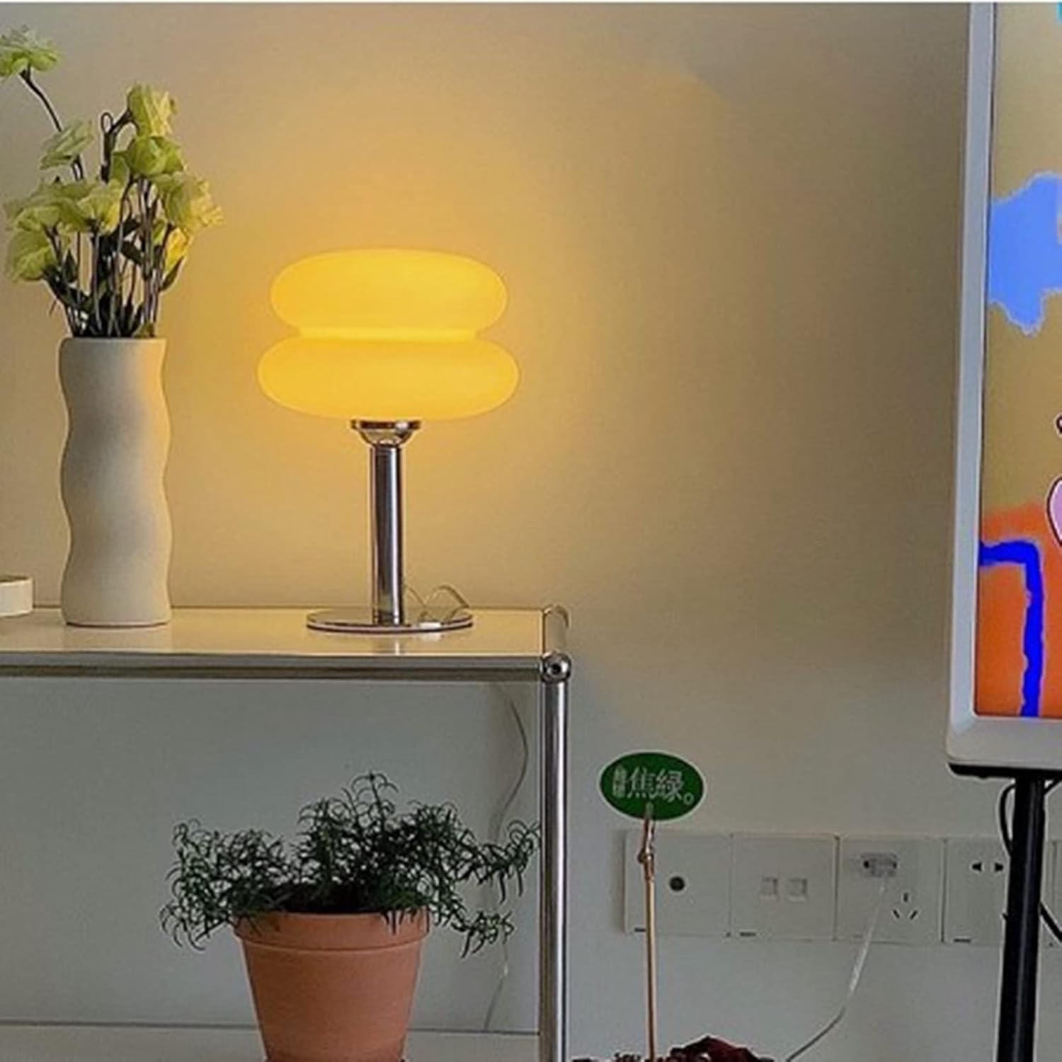 Eine gelbe, doppelstöckige Tischlampe steht auf einem weißen Regal neben einer Vase mit gelben Blumen und einer Topfpflanze.
