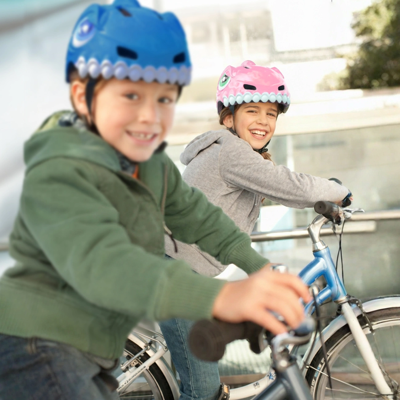 Due bambini sorridenti vanno in bicicletta all'aperto indossando divertenti caschi da dinosauro.