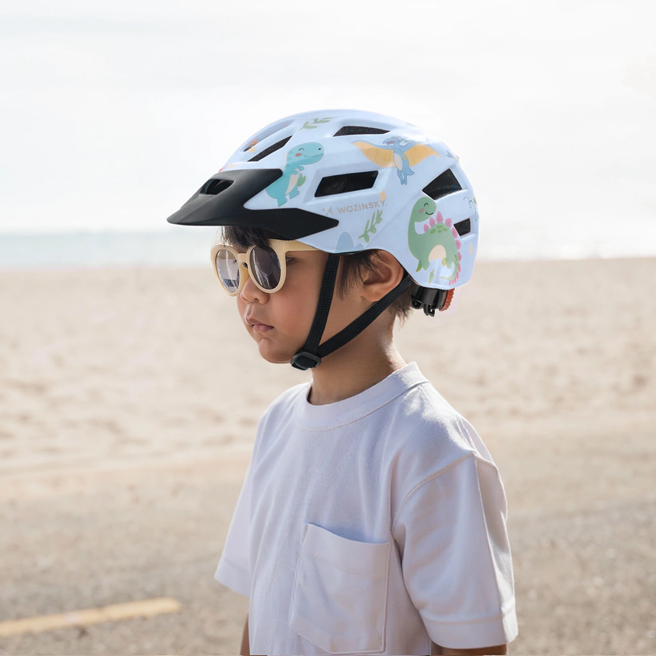 Un bambino con maglietta bianca, occhiali da sole e un casco da bicicletta con dinosauri.
