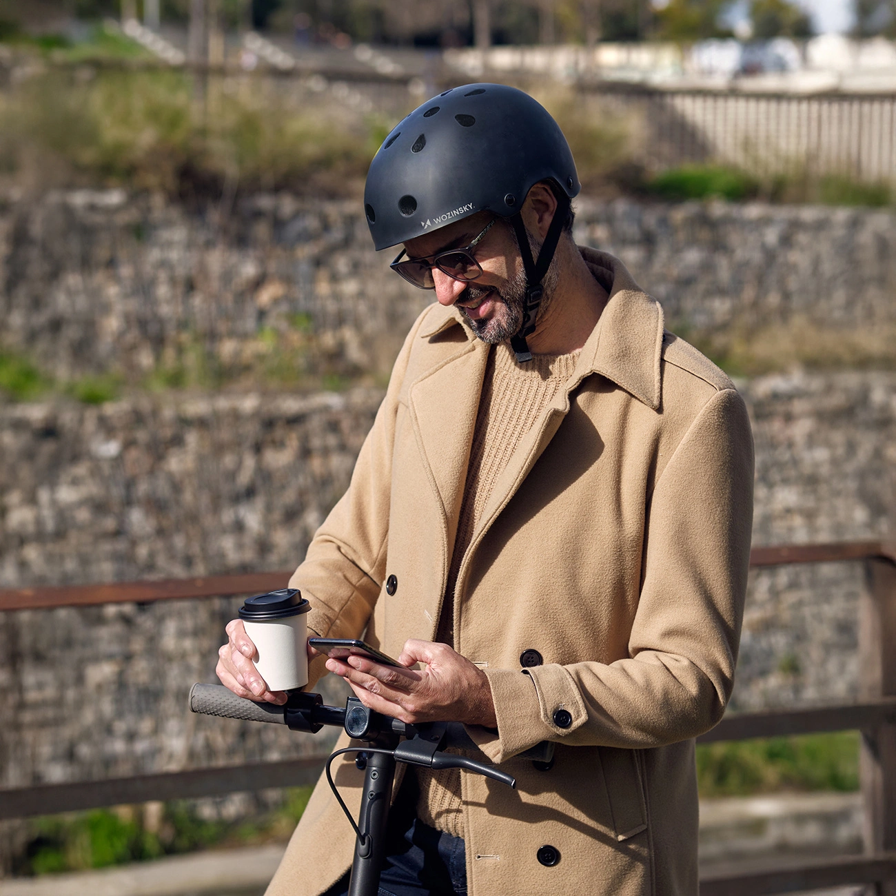 Un uomo con cappotto beige e casco tiene un caffè mentre usa lo smartphone su un monopattino.
