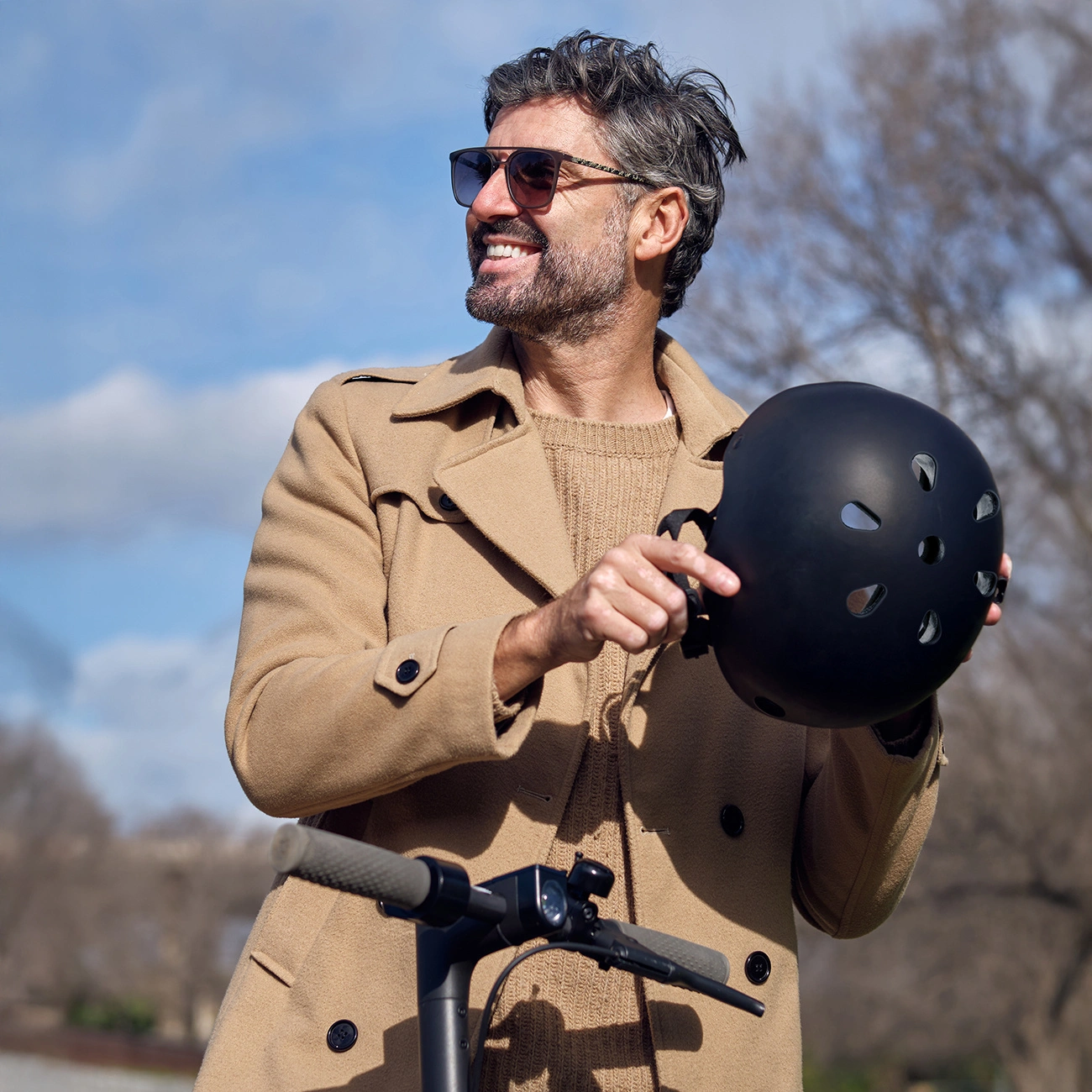 Uomo sorridente con cappotto beige e occhiali da sole tiene un casco nero vicino a un monopattino.