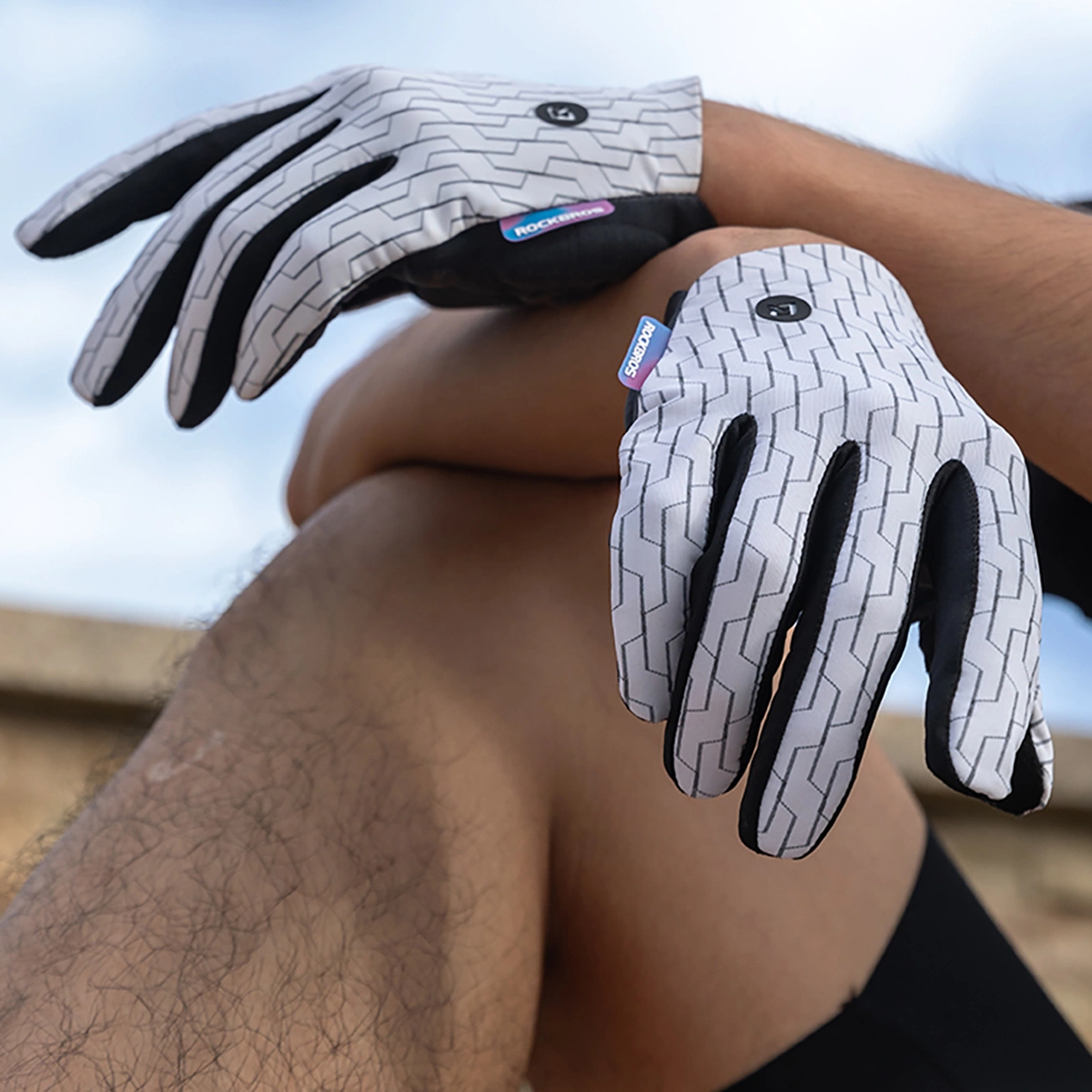 Persona che indossa guanti da ciclismo bianchi con motivo a mattoni grigi su cielo azzurro.