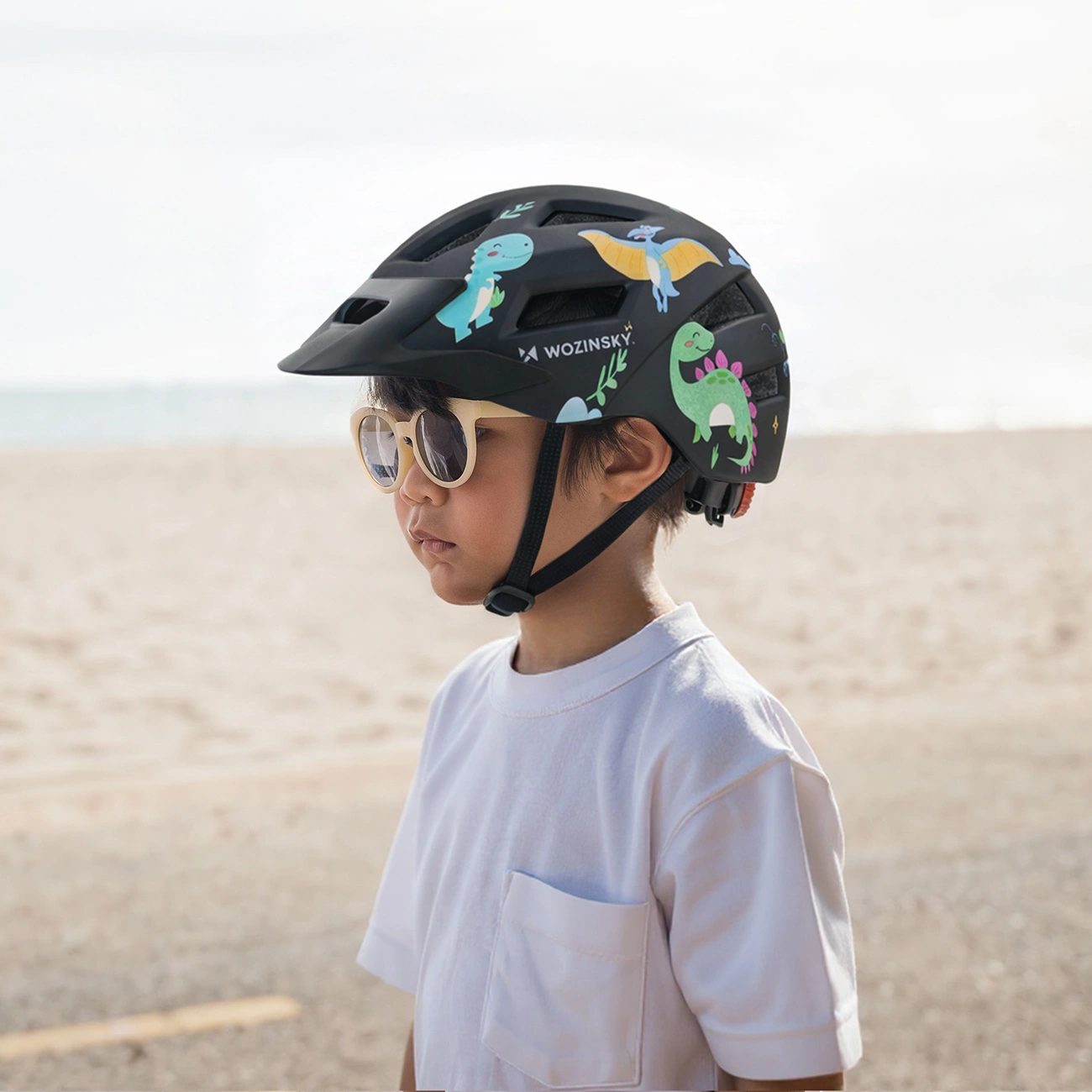 Un bambino con maglietta bianca, occhiali da sole e casco nero con adesivi di dinosauri si trova vicino a una spiaggia.