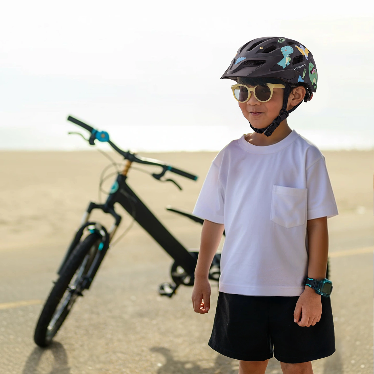 Un ragazzo con casco, occhiali da sole, maglietta bianca e pantaloncini neri si trova davanti a una bicicletta.