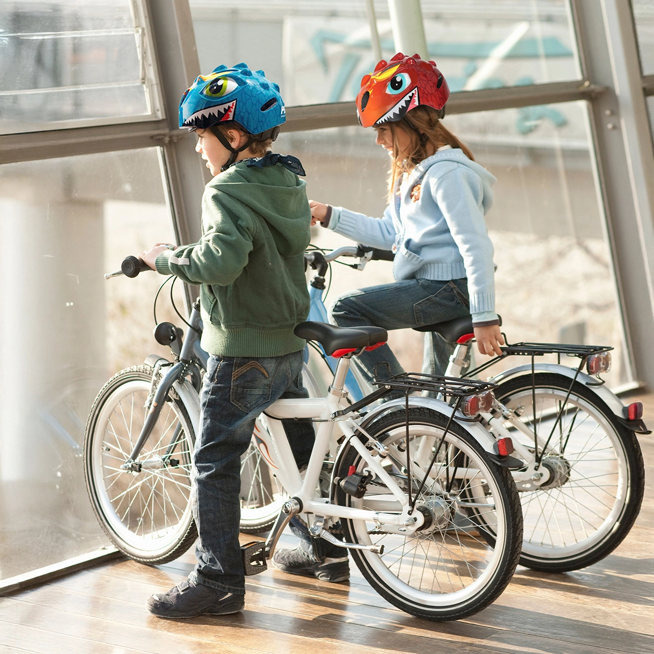 Due bambini con caschi da dinosauro sono seduti su biciclette bianca e blu davanti a una grande finestra.
