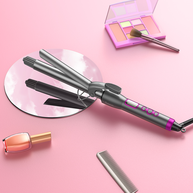 Ein grauer Lockenstab liegt auf einer rosa Oberfläche, umgeben von einem Make-up-Set, Nagellack und einem Kamm.