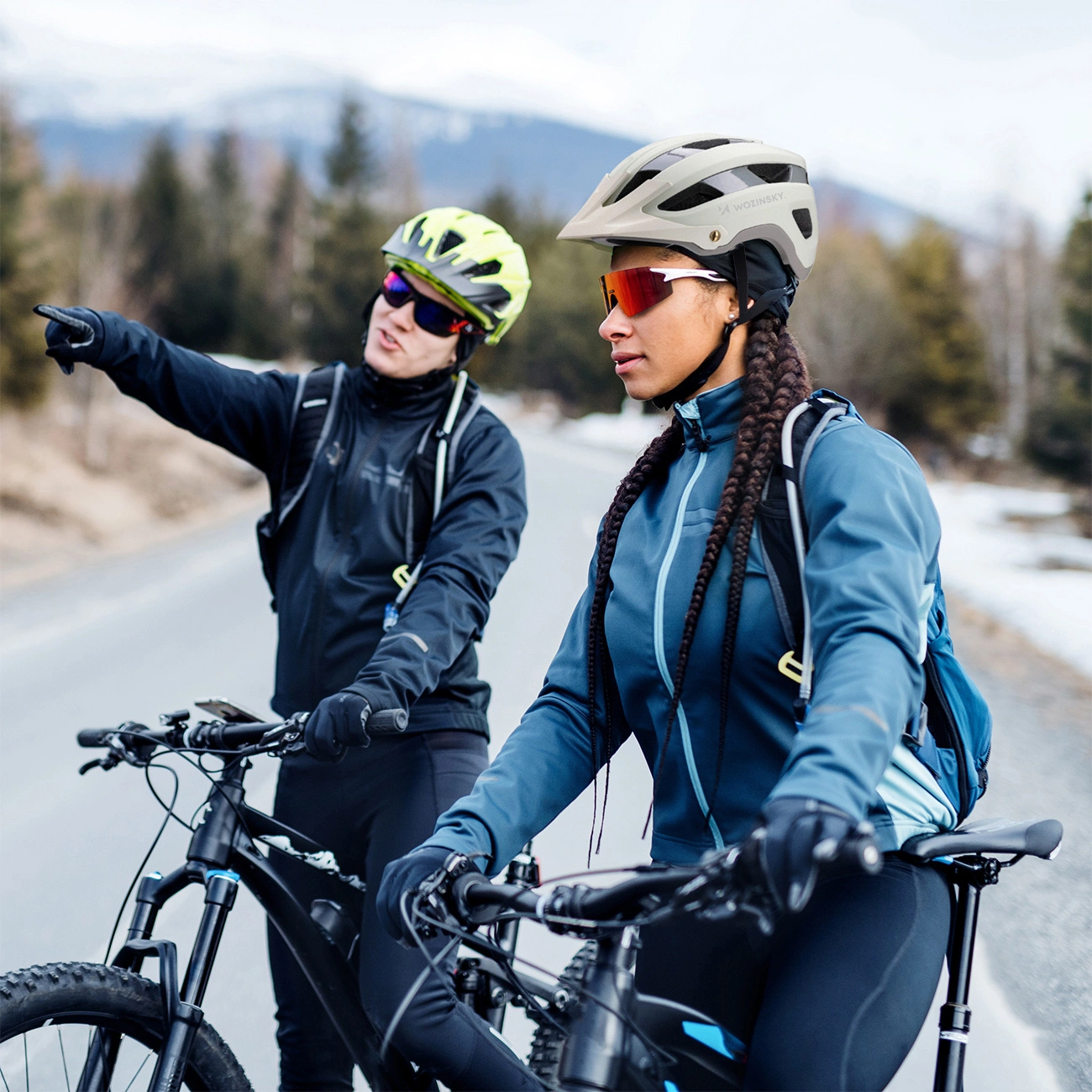 Due ciclisti con casco su una strada di montagna, uno indica qualcosa.