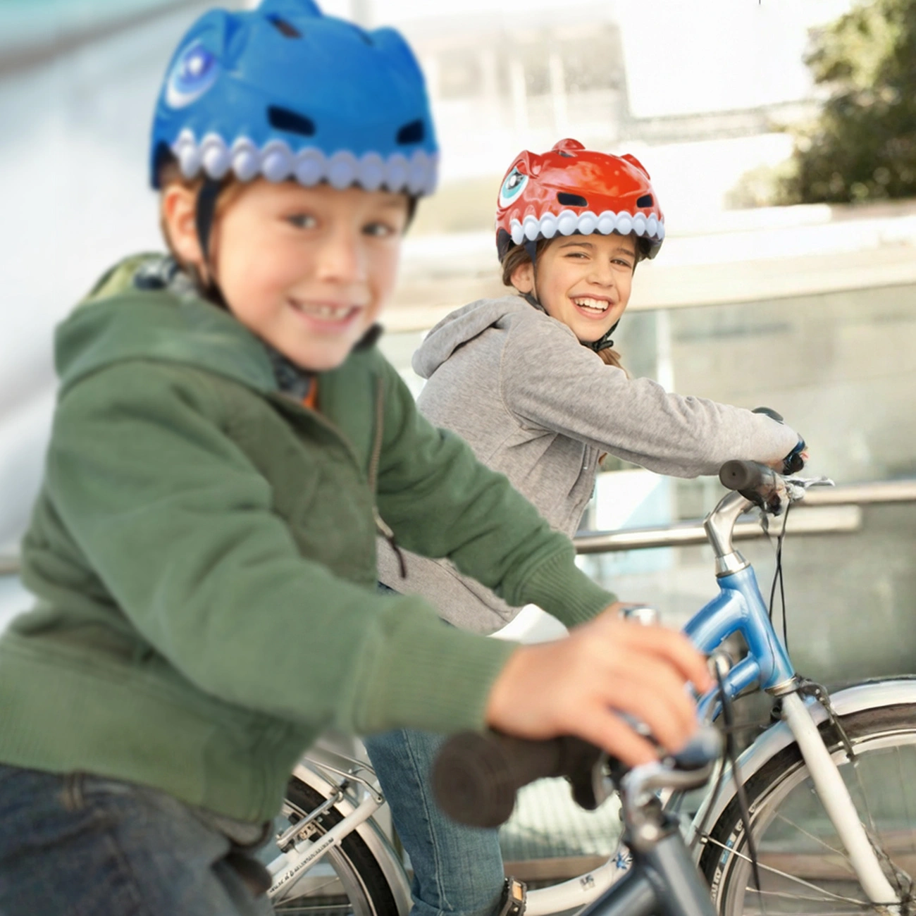 Due bambini sorridenti con caschi da bicicletta a forma di dinosauro vanno in bicicletta all'aperto.
