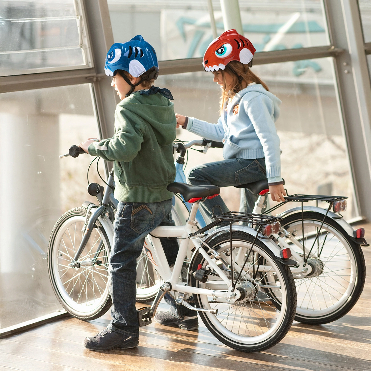 Due bambini con caschi di animali guidano biciclette vicino a una grande finestra.