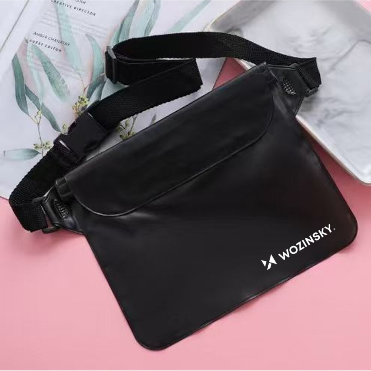 Una borsa a tracolla nera Wozinsky poggia su una superficie rosa con riviste e un vassoio bianco.