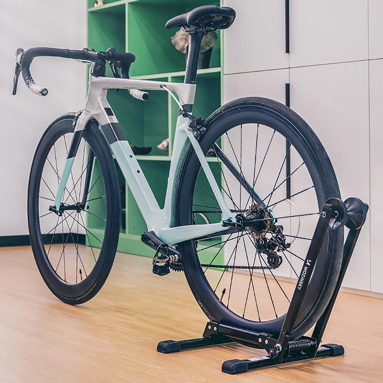 Una bici da corsa azzurra su un supporto nero in una stanza con scaffale verde e armadi bianchi.