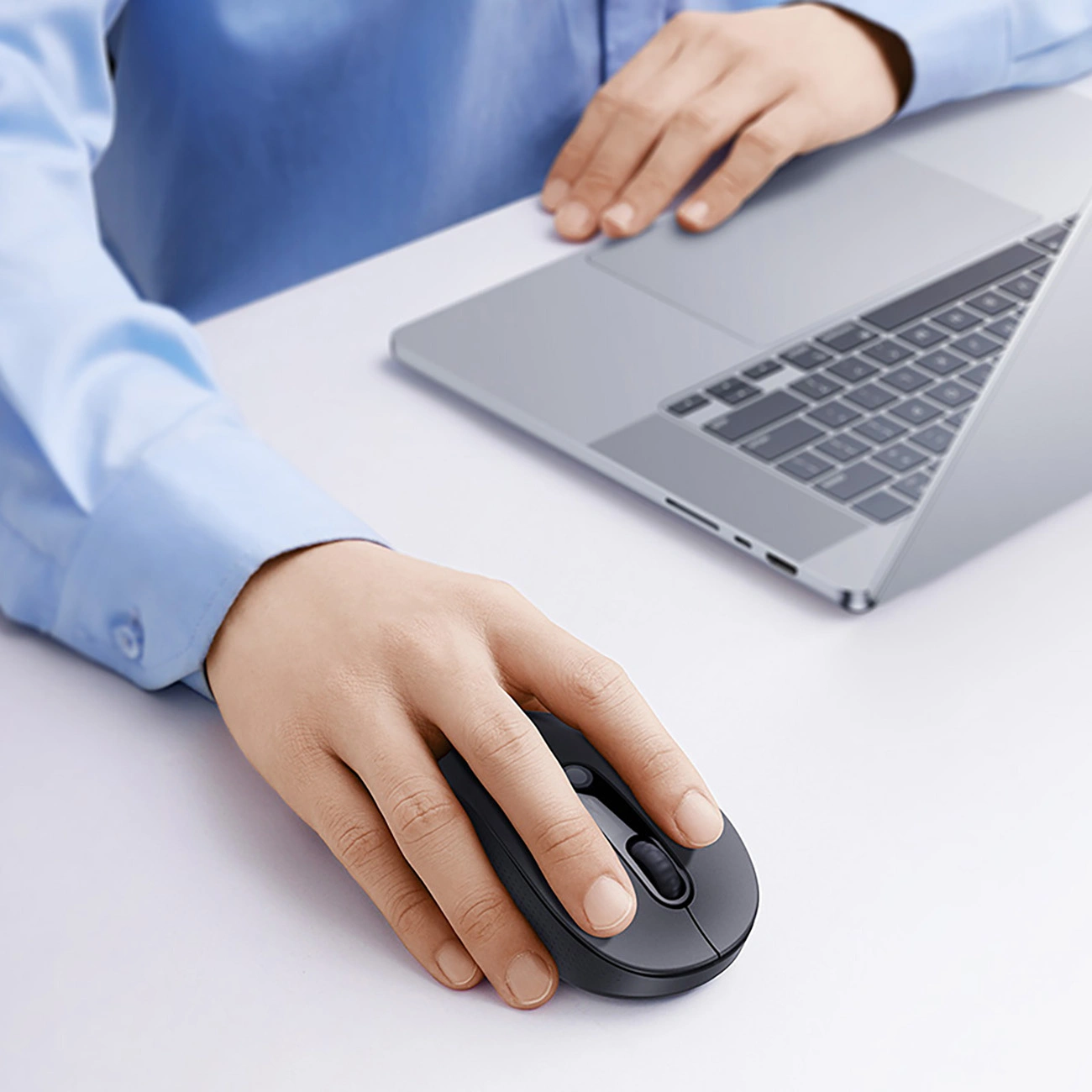 Persona con camicia blu usa un mouse grigio accanto a un laptop su una scrivania bianca.
