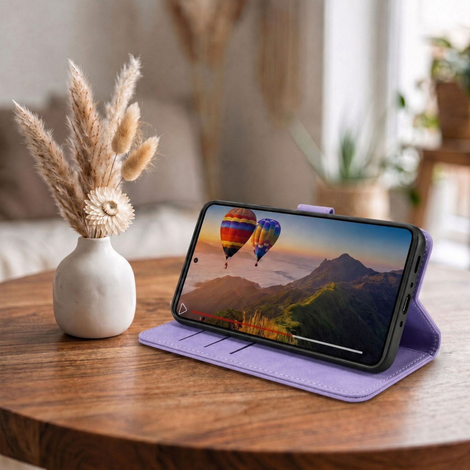 Lila Handyhülle mit aufgestelltem Smartphone auf Holztisch, daneben eine weiße Vase mit Trockenblumen.
