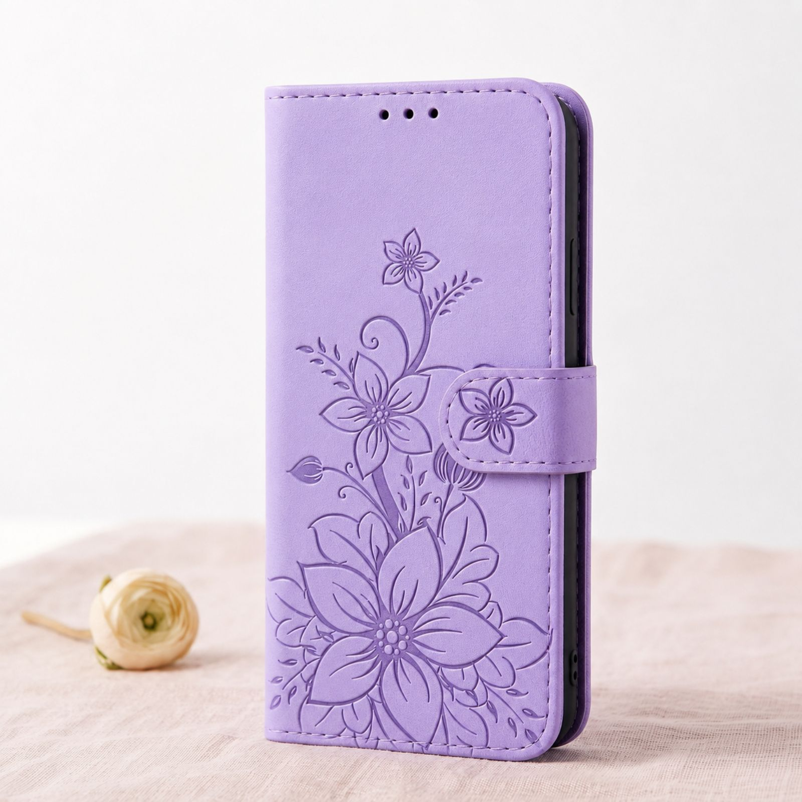 Eine lila Lederhandyhülle mit geprägten Blumenmustern steht aufrecht auf einem hellrosa Tuch neben einer weißen Blume.