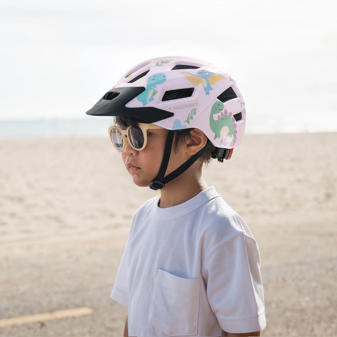 Un bambino con maglietta bianca indossa un casco da bici rosa con dinosauri e occhiali da sole.