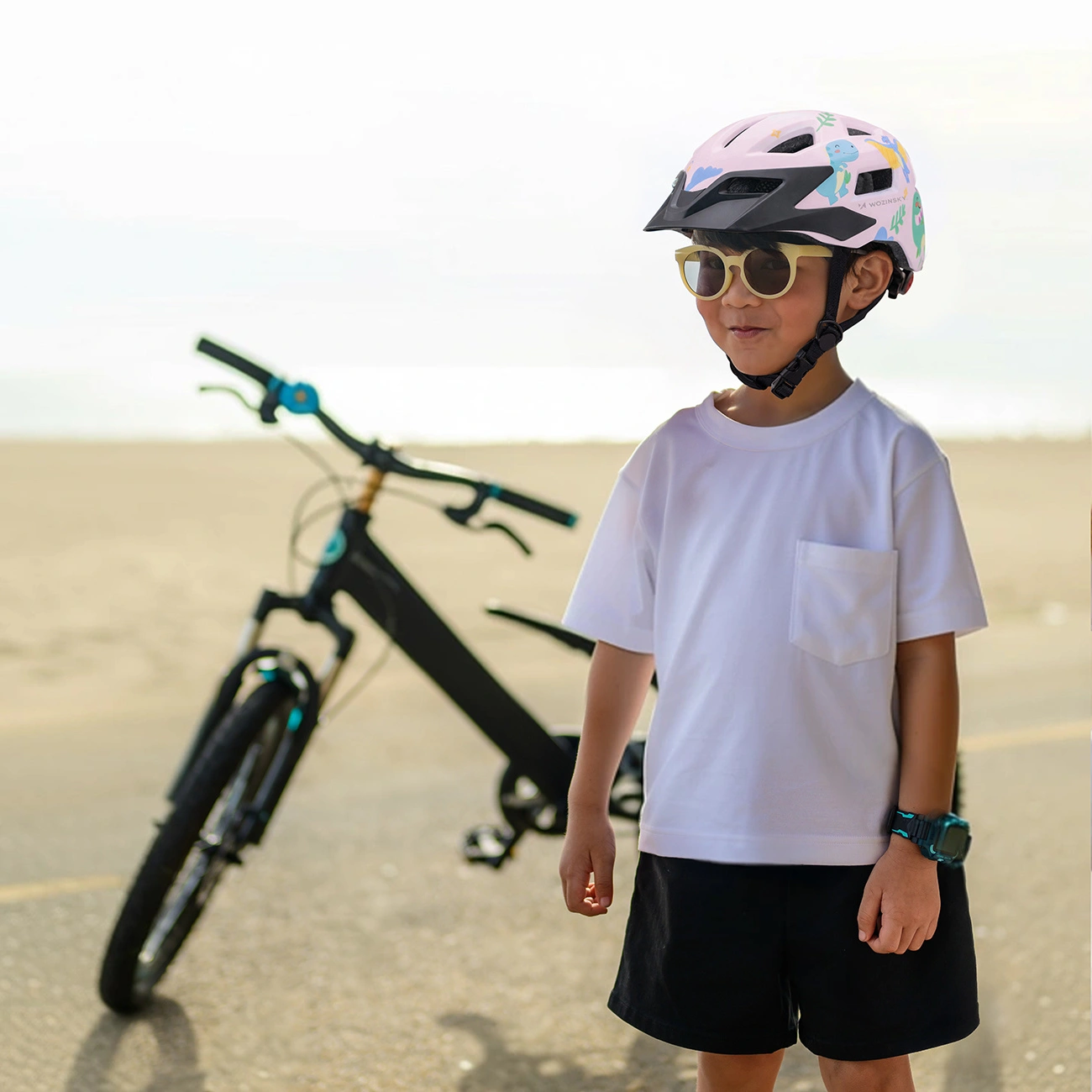 Un ragazzo in maglietta bianca, casco colorato e occhiali da sole è davanti a una bici.