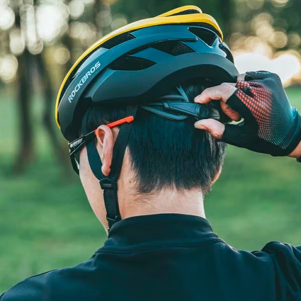 Un uomo con casco nero e guanti da ciclismo regola il sottogola. Sfondo verde naturale.