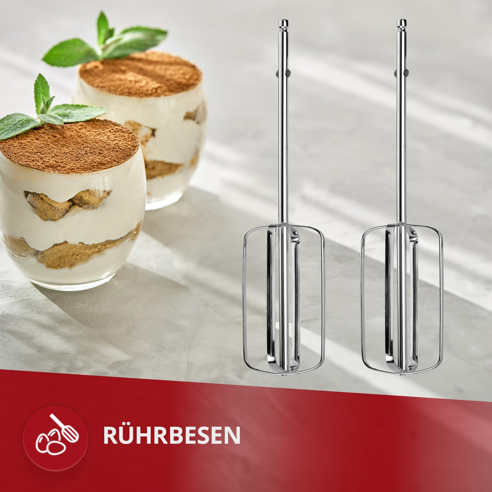 Zwei silberne Rührbesen stehen auf einer weißen Oberfläche neben zwei Gläsern Tiramisu; unten ein roter Balken mit Text.