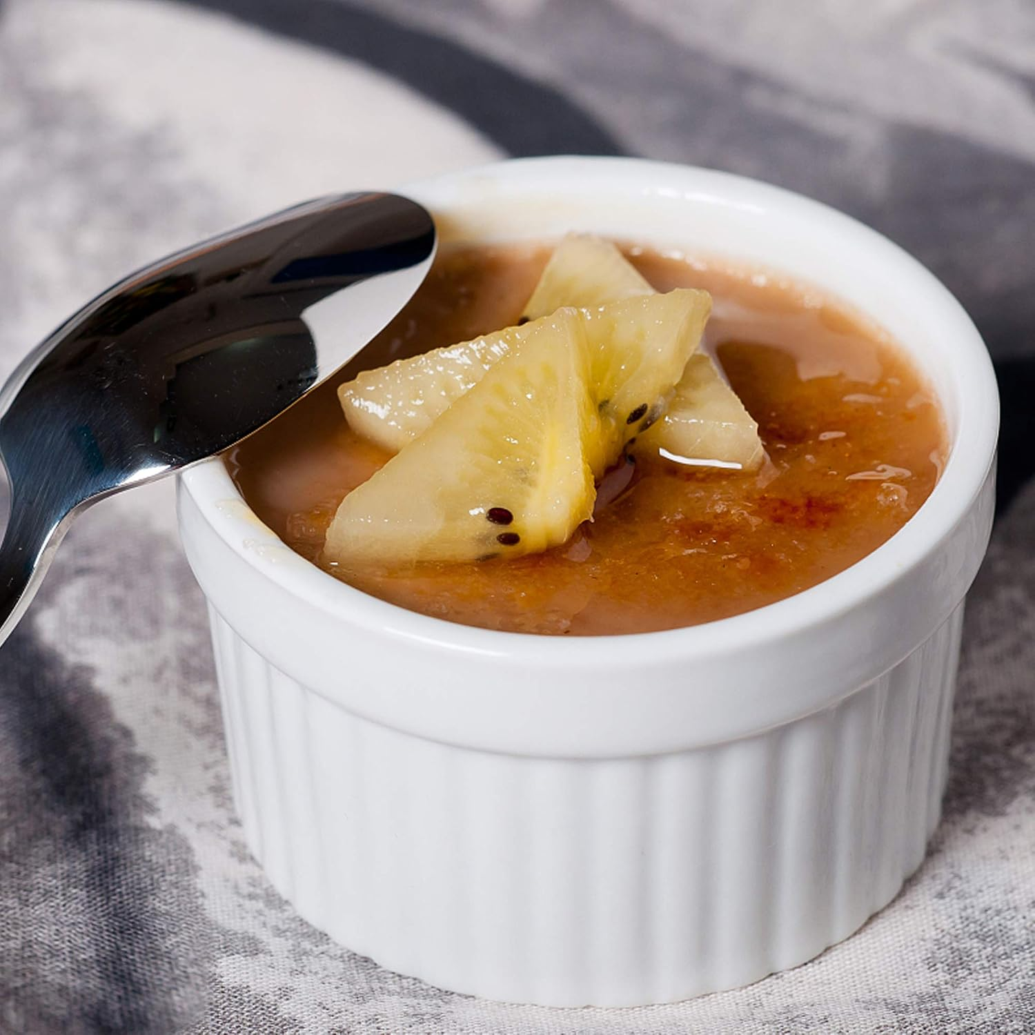 Eine weiße Schale mit Karamelldessert, gekrönt mit gelben Kiwischeiben und einem Löffel am Rand.