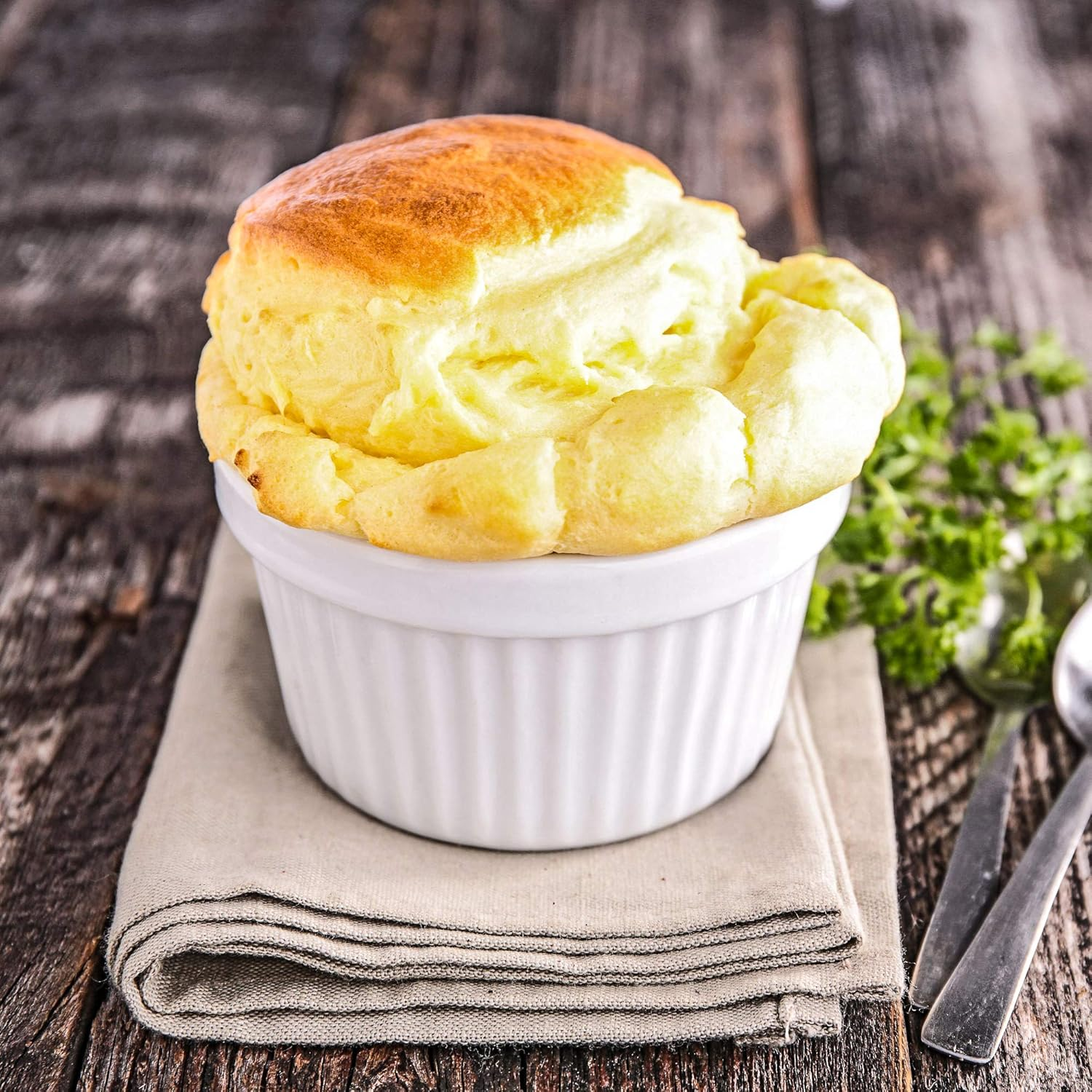 Ein goldbraun gebackener Käsesoufflé in einer weißen Auflaufform auf einer Serviette auf einem rustikalen Holztisch.