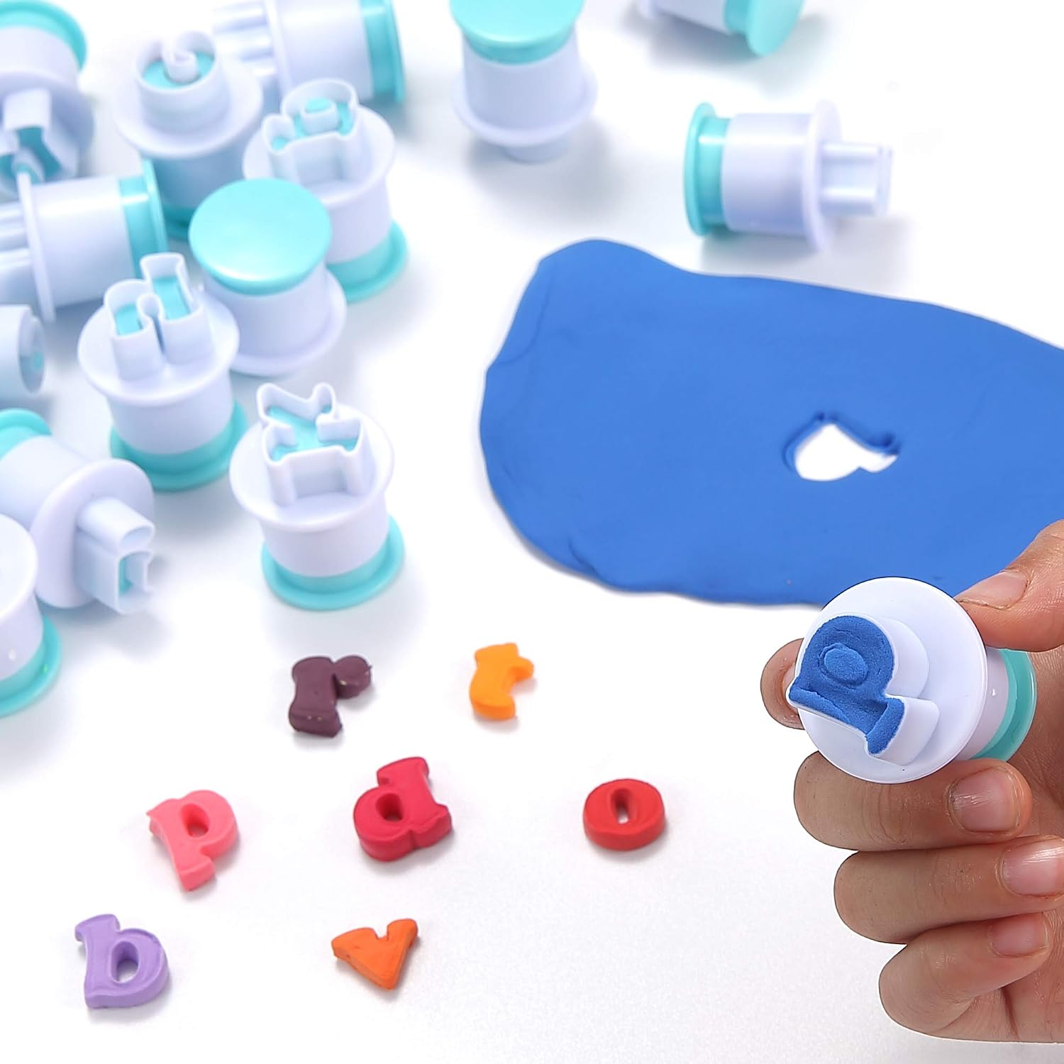 Hand hält Buchstabenausstecher über blauer Knete, daneben weitere Ausstecher und geformte Buchstaben.